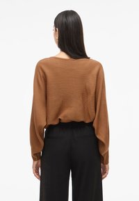 Brown ribbed sweater with wide sleeves, curved hem, and textured fabric. Paired with black trousers featuring subtle back pockets.