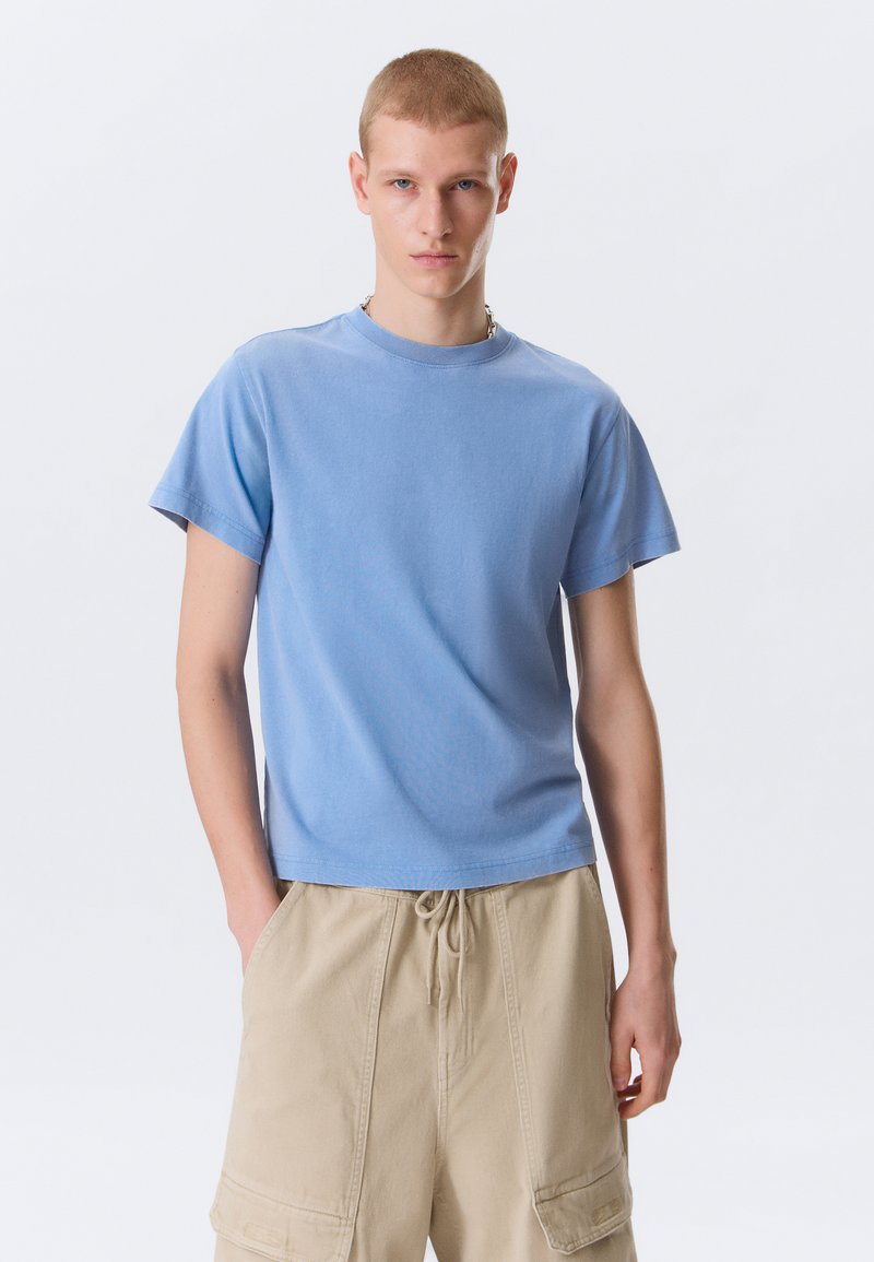 Junger Mann steht mit den Händen in den Taschen beigefarbener Cargo-Shorts und trägt ein schlichtes hellblaues kurzärmeliges T-Shirt vor hellem Hintergrund.