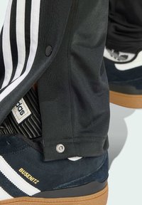 Adidas Busenitz sneakers i marinblå mocka med vita ränder och guld text, utrustade med en texturerad sula. Svarta träningsbyxor med tryckknappsstängning.