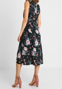 Vestito a stampa floreale con sfondo verde scuro, design senza maniche, gonna a lunghezza ginocchio e dettaglio con apertura a chiave sul retro. Abbinato a sandali con tacco rosa.