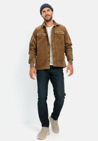 Bruine corduroy jas met knoopzakken op de borst, gecombineerd met donkerblauwe jeans en beige sneakers, tegen een effen witte achtergrond.