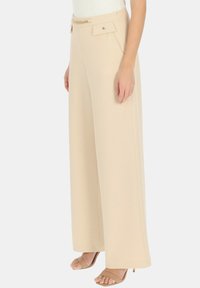 Beżowe spodnie o szerokich nogawkach z gładką fakturą, z przednimi kieszeniami, paskiem z łańcuszkiem oraz zapięciem na zamek zapewniającym bezpieczne dopasowanie.