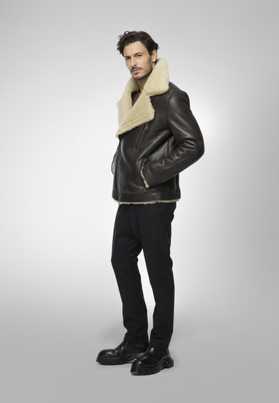 Giacca in shearling di pelle marrone con un ampio colletto, tasche con zip e una vestibilità rilassata, abbinata a pantaloni neri a gamba affusolata e stivali neri.