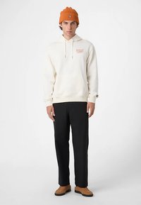 Krämvit hoodie med framsida grafik och dragsko, kombinerad med svarta byxor och bruna skor. Orange ribbad stickad mössa kompletterar looken.