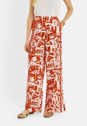 Mujer que lleva pantalones de talle alto y pierna ancha con estampado abstracto tropical en naranja y blanco, top sin mangas blanco, pulsera de brazalete dorado y sandalias beige.
