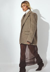 Femme marchant, portant un blazer beige duveteux sur un col roulé blanc et une jupe transparente brun foncé assortie à des bottes marron.