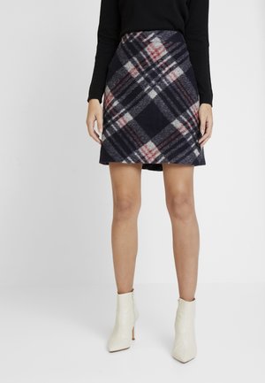 Woman wearing a navy, gray, and red plaid mini skirt with a black top and white heeled ankle boots standing against a white background.