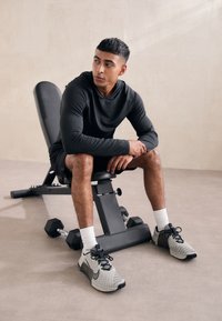 Schwarzes langärmliges Sportshirt, schwarze Shorts, graue Nike-Schuhe, sitzt auf einer grauen Gewichtsbank mit einer Kurzhantel in der Nähe. Glatte Bodenbeschaffenheit.