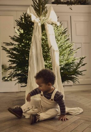 Un bambino seduto indossa una salopette di colore crema a pois con bottoni e una maglietta a maniche lunghe a righe scure, vicino a un albero di Natale verde.