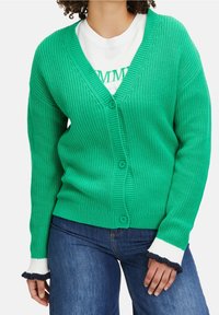 Žena v zeleném pleteném kardiganu na knoflíky s bílými a černými volánkovými manžetami, pod kterým má bílé tričko a modré džíny.