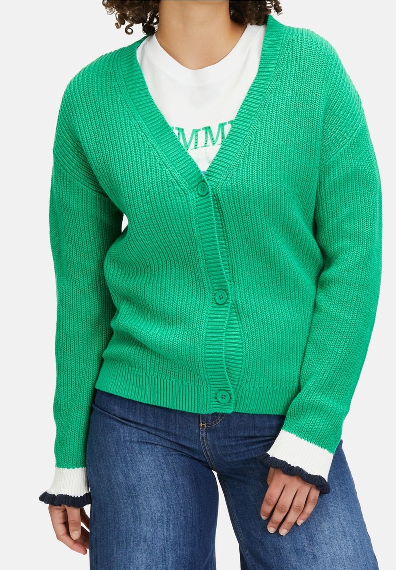Žena v zeleném pleteném kardiganu na knoflíky s bílými a černými volánkovými manžetami, pod kterým má bílé tričko a modré džíny.