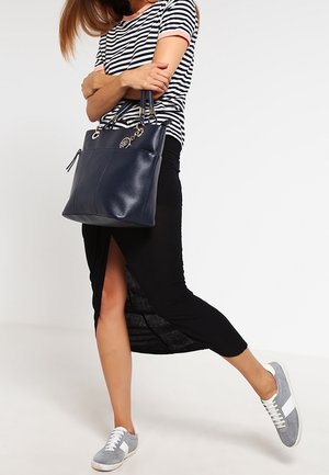 Mujer con camisa a rayas, falda negra con abertura, zapatillas grises, sosteniendo un bolso azul marino con los brazos cruzados.