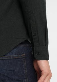 Dunkelgrünes Hemd mit Knopfleiste aus strukturiertem Stoff; verfügt über lange Ärmel, geknöpfte Manschetten und einen abgerundeten Saum. Kombiniert mit dunklen Denim-Jeans.