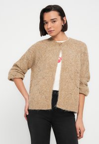 Νέα γυναίκα με κοντά σκούρα μαλλιά, φοράει μπεζ πλεκτή ζακέτα πάνω από λευκό πουκάμισο και μαύρα τζιν, στέκεται μπροστά σε απλό λευκό φόντο.