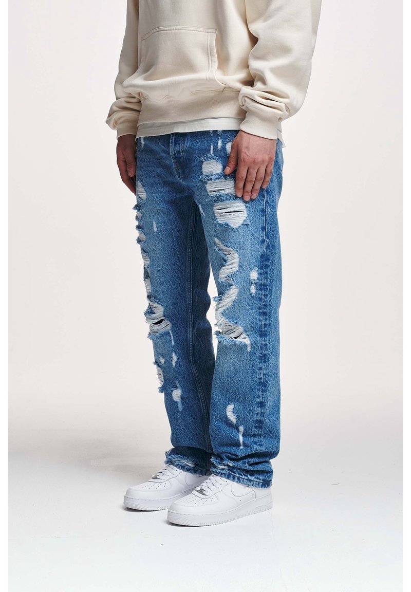 Homme portant un jean bleu déchiré, un sweat à capuche beige et des baskets blanches, debout devant un fond uni.