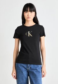Melna kokvilnas T-kreklu ar zelta "ck" logotipu vid�ū, īsām piedurknēm un klasisku apaļu kakla izgriezumu, kopā ar ziliem džinsiem.