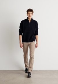 Un pull noir à côtes avec un col zippé mi-hauteur, assorti à un pantalon beige clair et des chaussures noires brillantes, le tout sur un fond neutre.