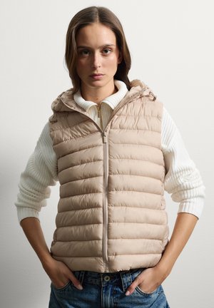 Gilet trapuntato beige con cappuccio, caratterizzato da una texture trapuntata e una zip frontale, indossato sopra un maglione a costine bianco. Vestibilità casual.