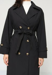 Manteau noir à double boutonnage, boutons oro, ceinture à nouer et manches larges. Tissu lisse avec un col classique.