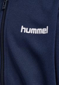 Tissu bleu marine avec le texte du logo « hummel » en blanc imprimé près d'une fermeture éclair noire sur un vêtement.
