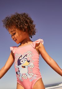 Rožinės maudymosi kostiumėliai su raukšlėtais petnešėliais, ant kurių pavaizduotas animacinis šuo, apsuptas gėlių. Lygus audinys, aptempianti forma, tinkami vandens veikloms.