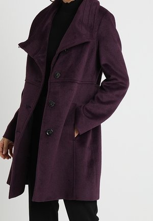 Femme portant un manteau en laine violet foncé boutonné avec un grand col, une main dans la poche, sur une tenue noire, sur un fond uni.