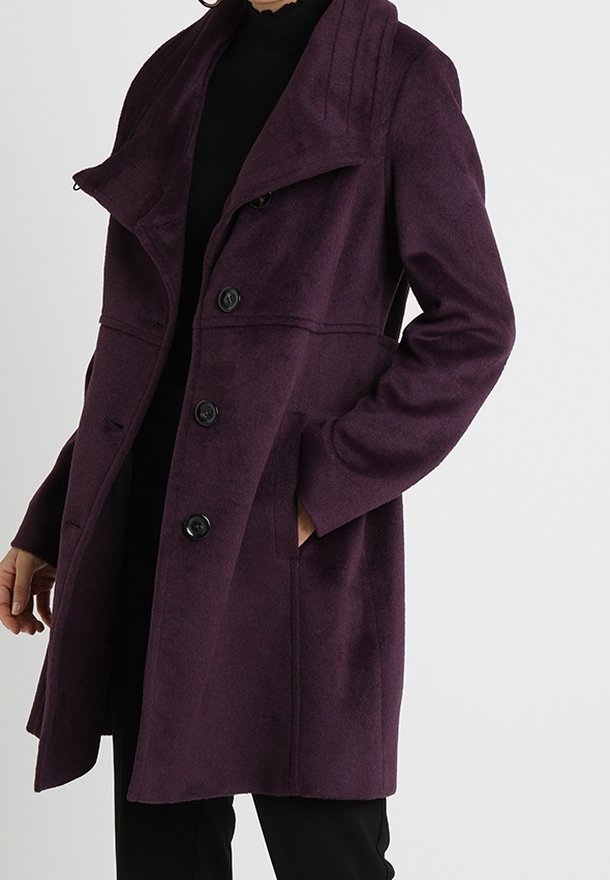 Femme portant un manteau en laine violet foncé boutonné avec un grand col, une main dans la poche, sur une tenue noire, sur un fond uni.