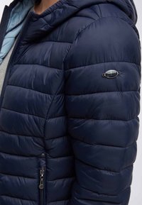 Dunkelblaue Steppjacke mit Reißverschluss, gekennzeichnet durch gesteppte Bereiche, eine Kapuze und ein kleines Logo-Detail am linken Arm.