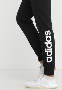 Zwarte sweatpants met een opvallend wit "adidas" logo aan de linkerkant, gecombineerd met witte mesh sportschoenen.