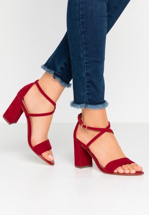 Røde ruskinds højhælede sandaler med blokhæle, åben tå og krydspåførte ankelremme. Hælen er cirka 7,5 cm høj.