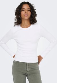 Λευκό μπλουζάκι μακρυμάνικο ριμπ με στρογγυλή λαιμόκοψη, συνδυασμένο με παντελόνι λαδί, παρουσιάζοντας εφαρμοστή σιλουέτα και λεία υφή.