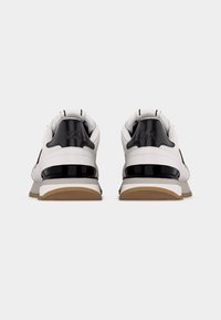 Paire de baskets blanches avec des accents noirs sur le talon et des semelles marron, vues de dos sur un fond blanc uni.