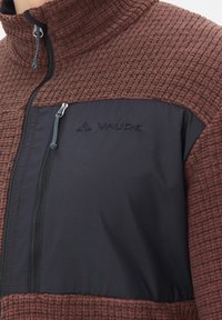 Giacca in pile con un esterno testurizzato marrone e un pannello nero liscio; presenta una tasca con zip e logo ricamato.
