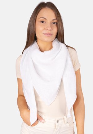 Junge Frau trägt einen großen weißen texturierten Schal über einem beigen kurzärmeligen Oberteil, mit einer Hand in der Tasche, vor einem schlichten Hintergrund.