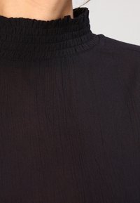 Blouse noire avec un col haut froncé, tissu léger à texture subtile, et détails de coutures apparentes.