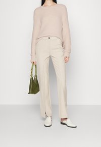 Kobieta w różowym, puszystym swetrze, beżowych spodniach z przodem z rozcięciami, białych lordach, trzymająca zieloną zamszową torebkę, stojąca na białym tle.