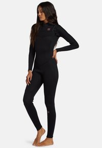 Muta in neoprene nera con maniche lunghe, design aderente, cuciture flatlock e dettaglio con zip sulla spalla, adatta per attività acquatiche.