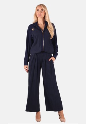 Marineblaues Zweiteiler-Outfit mit einer Zip-Jacke und einem runden Patch sowie einer weit geschnittenen Hose mit Falten. Glatter Stoff, elegantes Design.