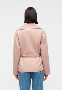 Γυναίκα που φαίνεται από πίσω, φοράει ανοιχτό ροζ παλτό με ζώνη και μπλε τζιν, πάνω σε μια απλή λευκή φόντο.