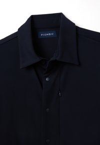 Camicia blu scuro con bottoni e collo strutturato, caratterizzata da una texture liscia e un piccolo dettaglio di tasca sul davanti. Etichetta del marchio visibile.