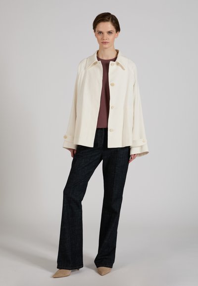 Donna che indossa giacca crema con bottoni sopra top marrone, pantaloni larghi scuri e scarpe beige a punta, in piedi davanti a uno sfondo grigio semplice.