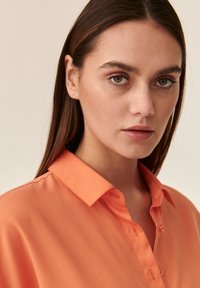 Camicia arancione con colletto rigido, texture liscia e leggero splendore, caratterizzata da bottoni piccoli e un design aderente.