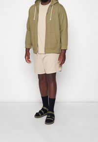 Hombre vestido con una sudadera con cremallera verde oliva, pantalones cortos beige, calcetines negros y sandalias verde oliva, de pie contra un fondo blanco.