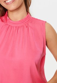 Top senza maniche di un brillante colore rosa, con un colletto plissettato alto e una texture liscia. Design semplice senza ulteriori motivi o dettagli.