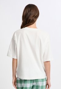 T-shirt z krótkim rękawem, okrągłym dekoltem, w kolorze off-white, wykonany z miękkiego materiału, zestawiony z zielonymi spodniami w kratę. Minimalistyczny design, luźny krój.