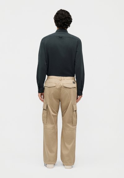 Άνδρας στέκεται πλάγια με μαύρο μακρυμάνικο πουκάμισο και μπεζ παντελόνι cargo με πίσω φάκελο και πλαϊνές τσέπες σε λευκό φόντο.