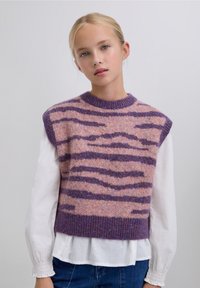 Suéter sin mangas de forro polar en rosa y púrpura con rayas de cebra. Cuello redondo y dobladillo acanalados. Llevado sobre una camisa blanca de manga larga.