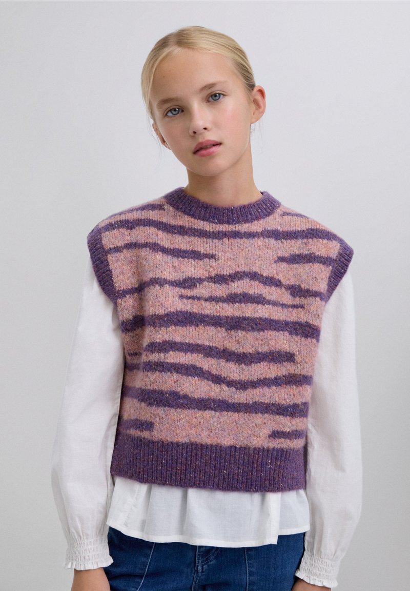 Suéter sin mangas de forro polar en rosa y púrpura con rayas de cebra. Cuello redondo y dobladillo acanalados. Llevado sobre una camisa blanca de manga larga.