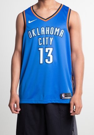 Persoon draagt een blauw Oklahoma City basketbalshirt met nummer 13 en zwarte korte broek, staand tegen een effen achtergrond.