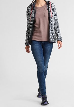 Kobieta nosząca wzorzystą kurtkę na zamek, mauve bluzkę z długim rękawem, niebieskie dżinsy i czarno-fioletowe trampki idzie do przodu.